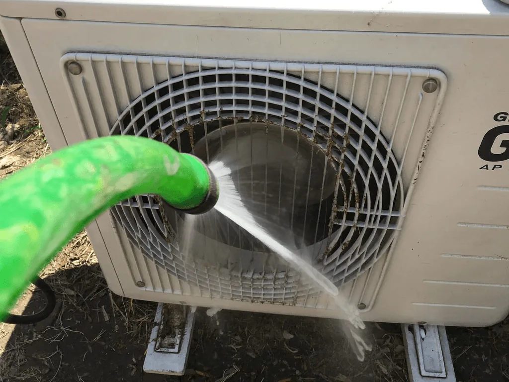 Green hose spraying water to rinse a dirty outdoor A/C condenser unit
