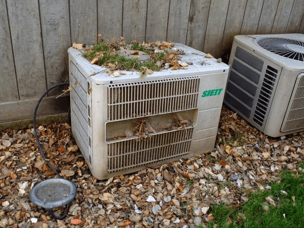 Neglected A/C condenser unit clogged with leaves and debris beside a fence